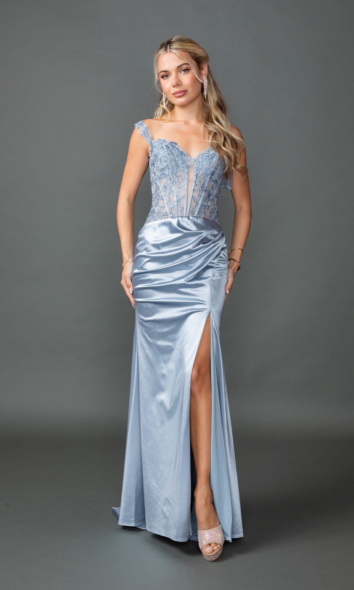 A woman stands against a gray background wearing the Dancing Queen 4536 long prom dress—a light blue, lace corset gown with a sweetheart neckline, thin straps, floor-length hem, and high slit—paired with open-toe heels.